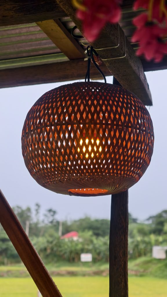 A close-up of a woven bamboo lantern glowing warm orange, with blurred flowers and greenery in the background.