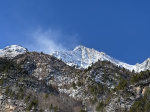 

Snow-dusted mountain peak under clear blue sky. Foreground features green, wooded slopes. The scene conveys a sense of tranquility and majesty.