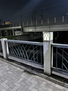 Nighttime view of a bridge with a glowing lamp post illuminating decorative railings, with an overpass and distant buildings in Shin-Chiba Park