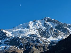 Snow-capped mountain peak under a clear blue sky with a crescent moon.