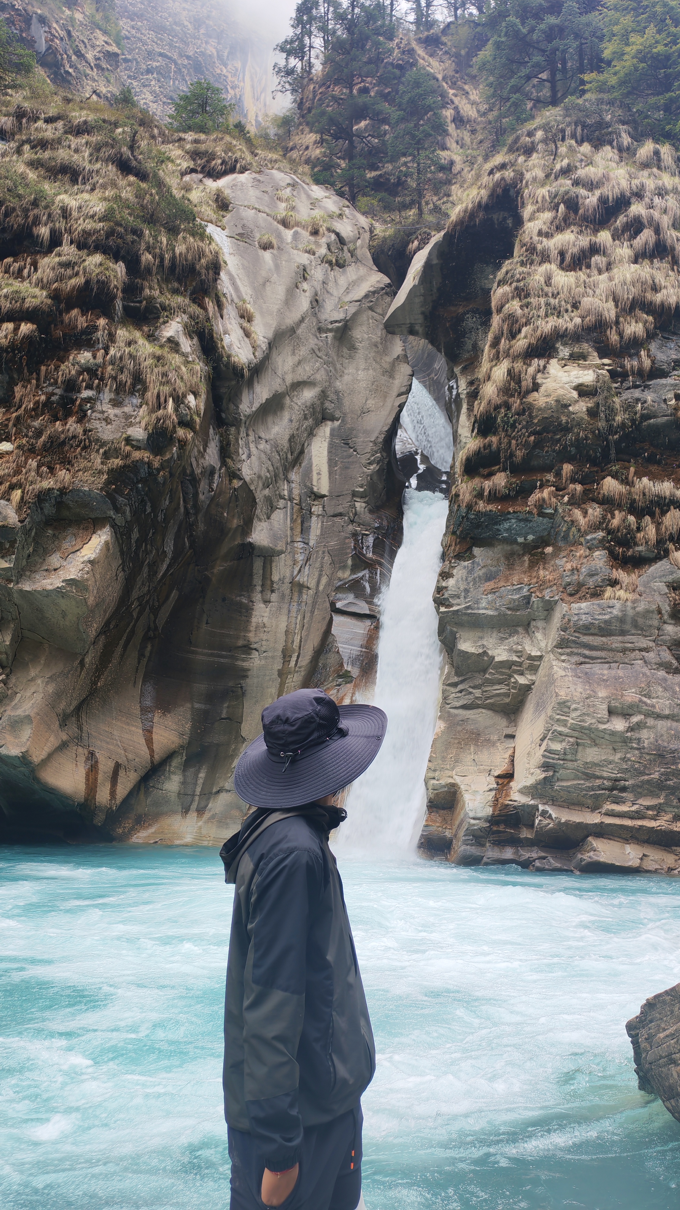 A person stands near a vibrant turquoise river, surrounded by towering rock formations and steep cliffs covered in sparse vegetation. The individual, dressed in a dark jacket and a wide-brimmed hat, gazes at a waterfall cascading down from a rock opening.