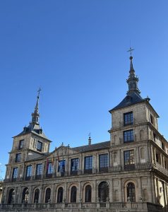 A grand historical building stands prominently against a clear blue sky. It features an intricate façade with multiple levels, large arched windows adorned with wrought iron balconies, and decorative stonework.