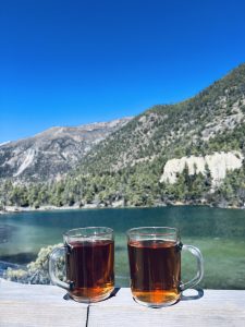 Two clear mugs of tea sit on a wooden ledge overlooking a serene turquoise lake and lush green mountains under a bright blue sky.
