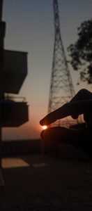 A hand appears to pinch the setting sun between two fingers, with silhouetted buildings and an electric tower against an orange and blue evening sky.