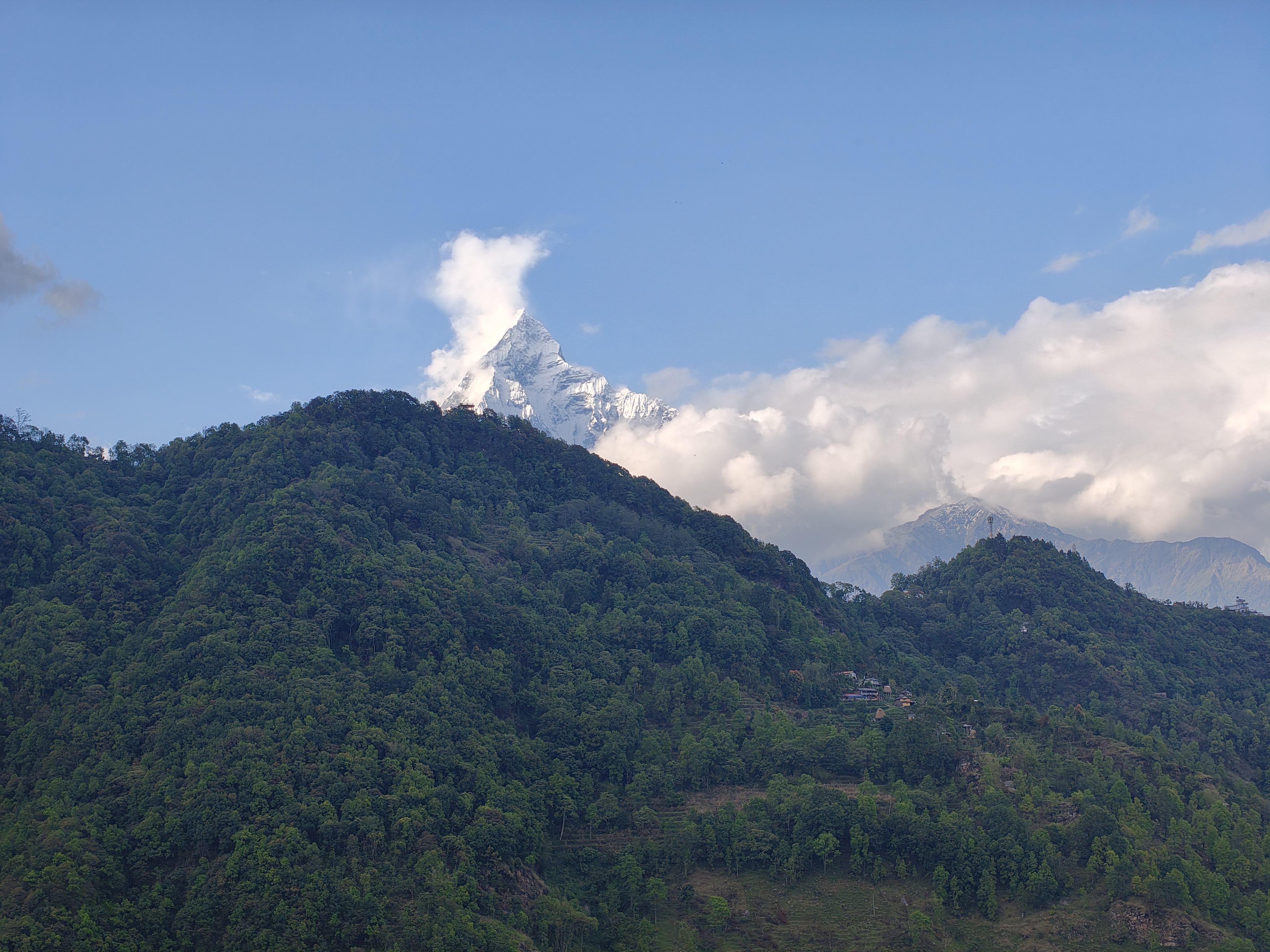 Snow-capped mountain peak emerging from clouds above lush green hills under a clear blue sky, portraying a serene and majestic landscape.