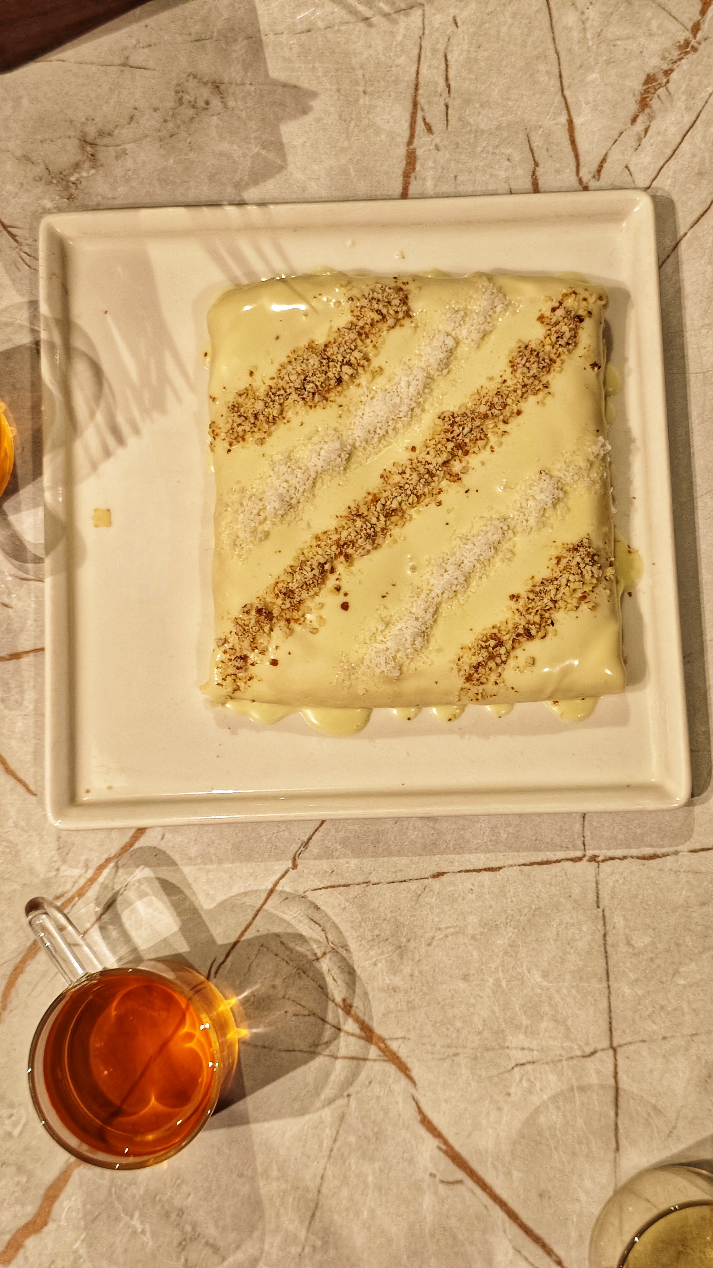 A square dessert is presented on a white plate, topped with a creamy layer and sprinkled with nuts and shredded coconut.