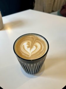 A glass cup of coffee with heart-shaped latte art on top, placed on a white table.