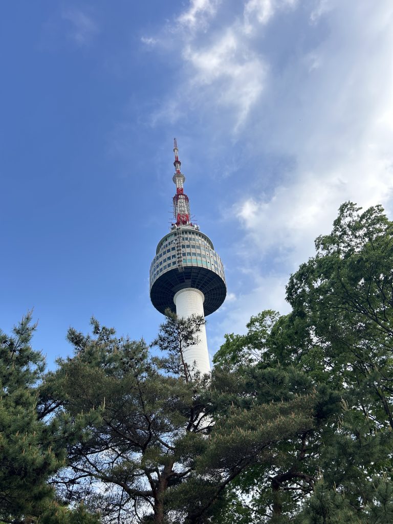Seoul Tower with its red antenna rises against a blue sky, framed by lush green trees at the base, evoking a sense of height and grandeur.
