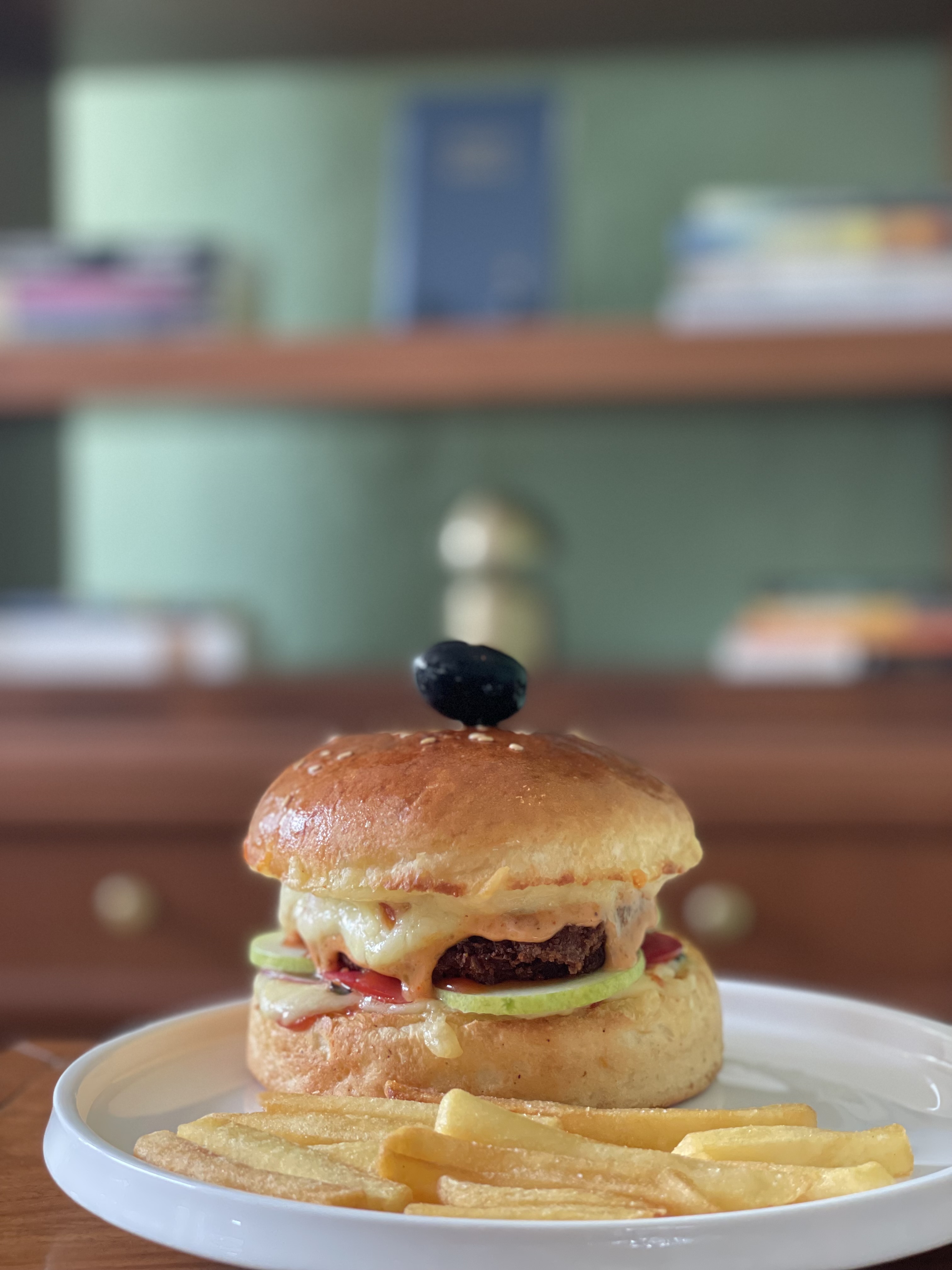 A close-up, eye-level shot of a juicy gourmet burger served on a white plate with a side of french fries. The burger features melted cheese, a dark patty, sliced cucumber, and a single black olive pinned to the top bun.
