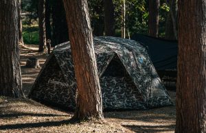 A camouflage-patterned tent is set up in a wooded area, surrounded by tall pine trees. The tent has a rounded top and is partially obscured by a tree trunk, suggesting a rustic, outdoorsy setting.