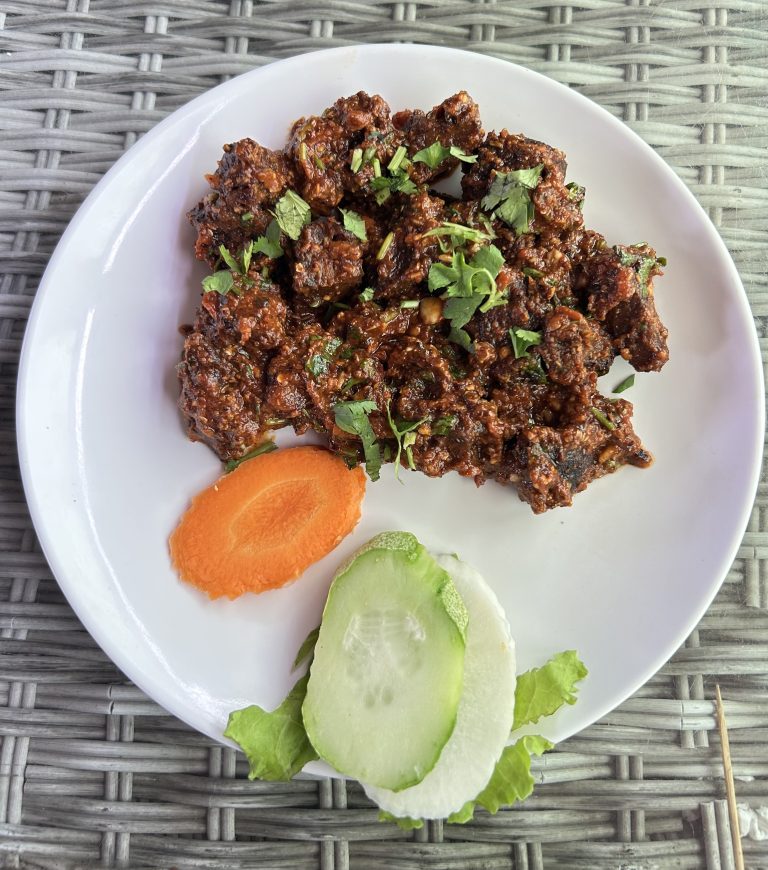 Spicy dish garnished with cilantro on a white plate, accompanied by sliced cucumber and carrot, on a woven textured surface. Vibrant and appetizing.