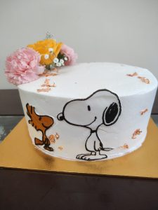 A cake with a minimalist Snoopy and Woodstock design is decorated with small flowers and golden details on a smooth white surface.