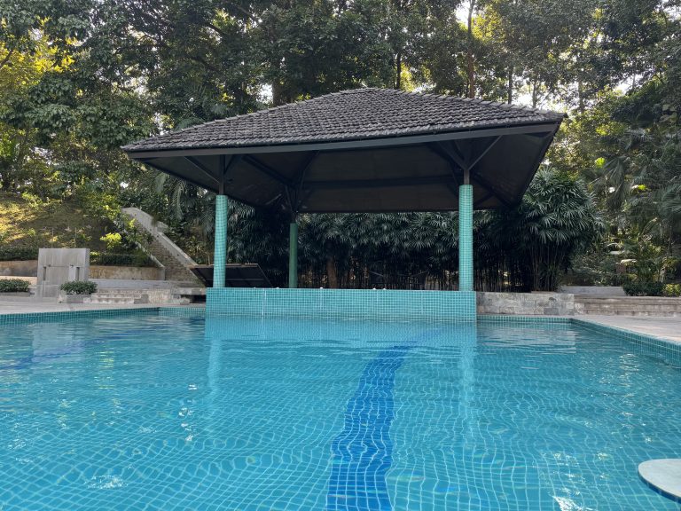 A clear blue swimming pool with turquoise mosaic tiles leads to an open-air pavilion under a dark tiled roof, surrounded by lush green tropical trees and foliage.