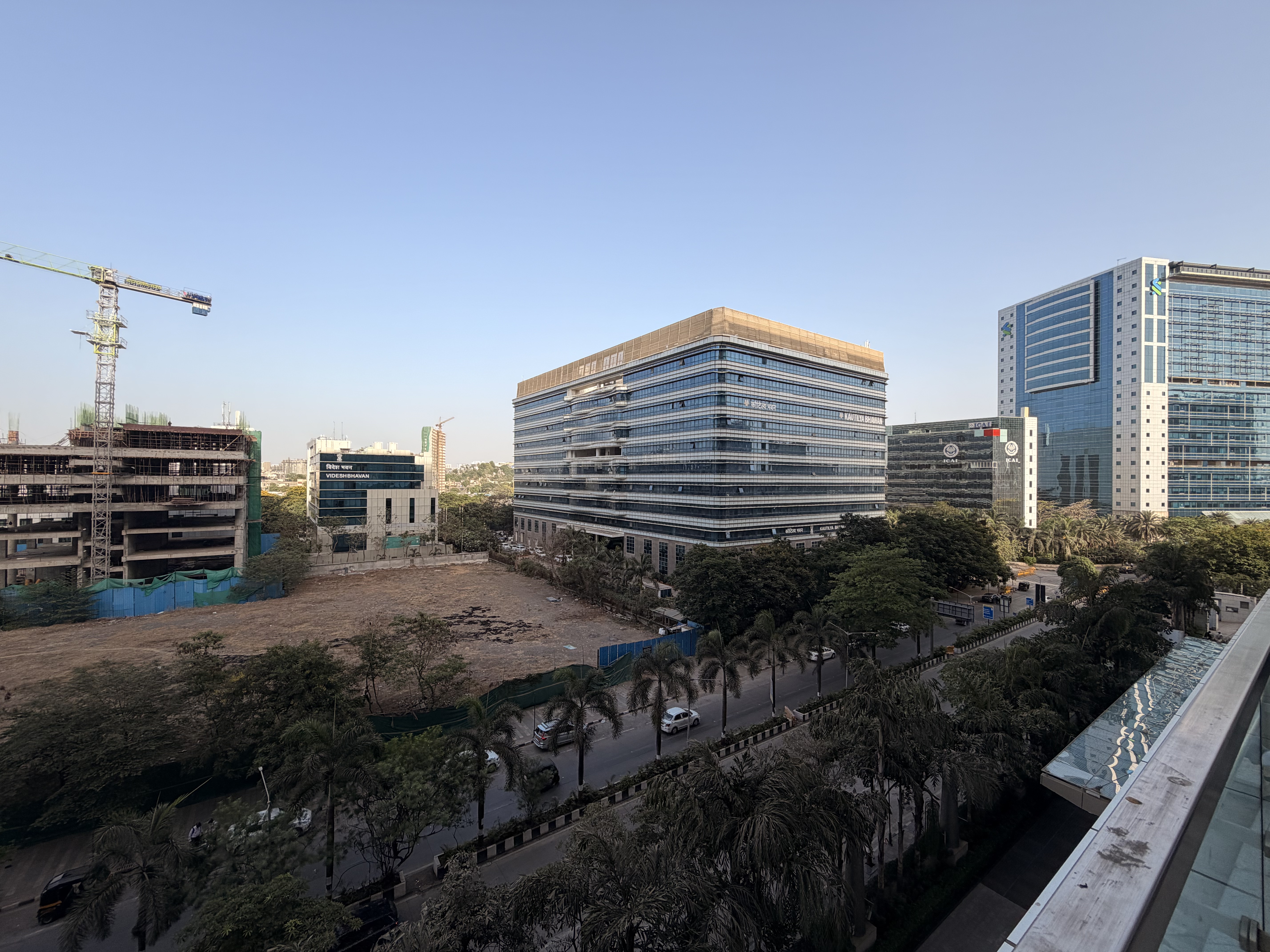 A panoramic view of a modern city skyline with glass office buildings, palm-lined streets, and ongoing construction. The scene captures urban development and a clear blue sky.