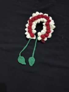 A crocheted floral garland with red and white flowers and green leaves is displayed on a black background, highlighting handcrafted artistry.