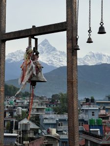 Bell with colorful cloths hangs in the foreground, a cityscape below. Snow-capped mountains rise majestically in the background under a clear sky.