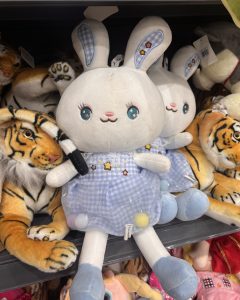 A cute plush bunny with wide teal eyes and white fur wears a blue checkered dress with stars and pom-poms