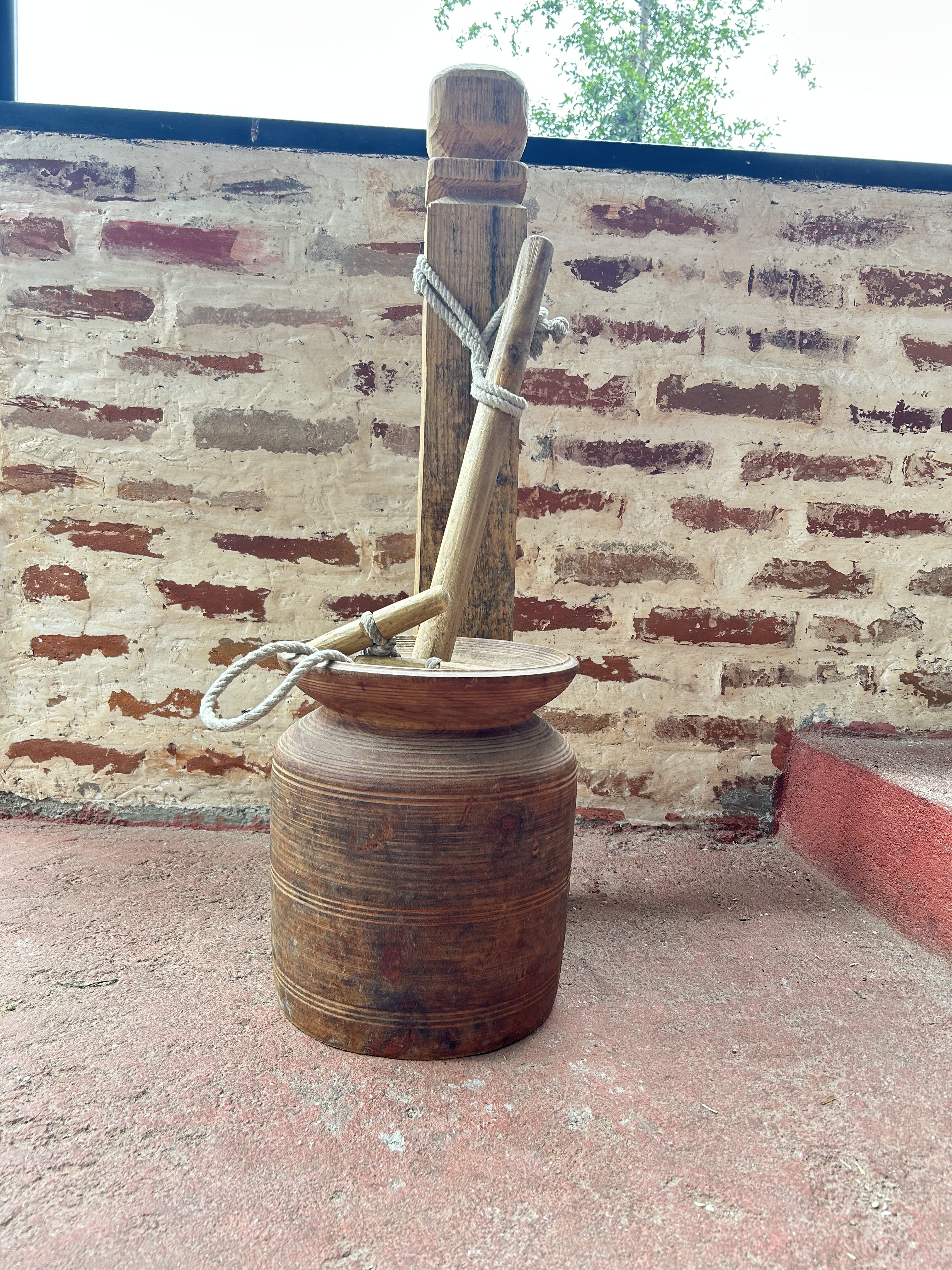A rustic wooden butter churn with a paddle leans against a textured, weathered brick wall.