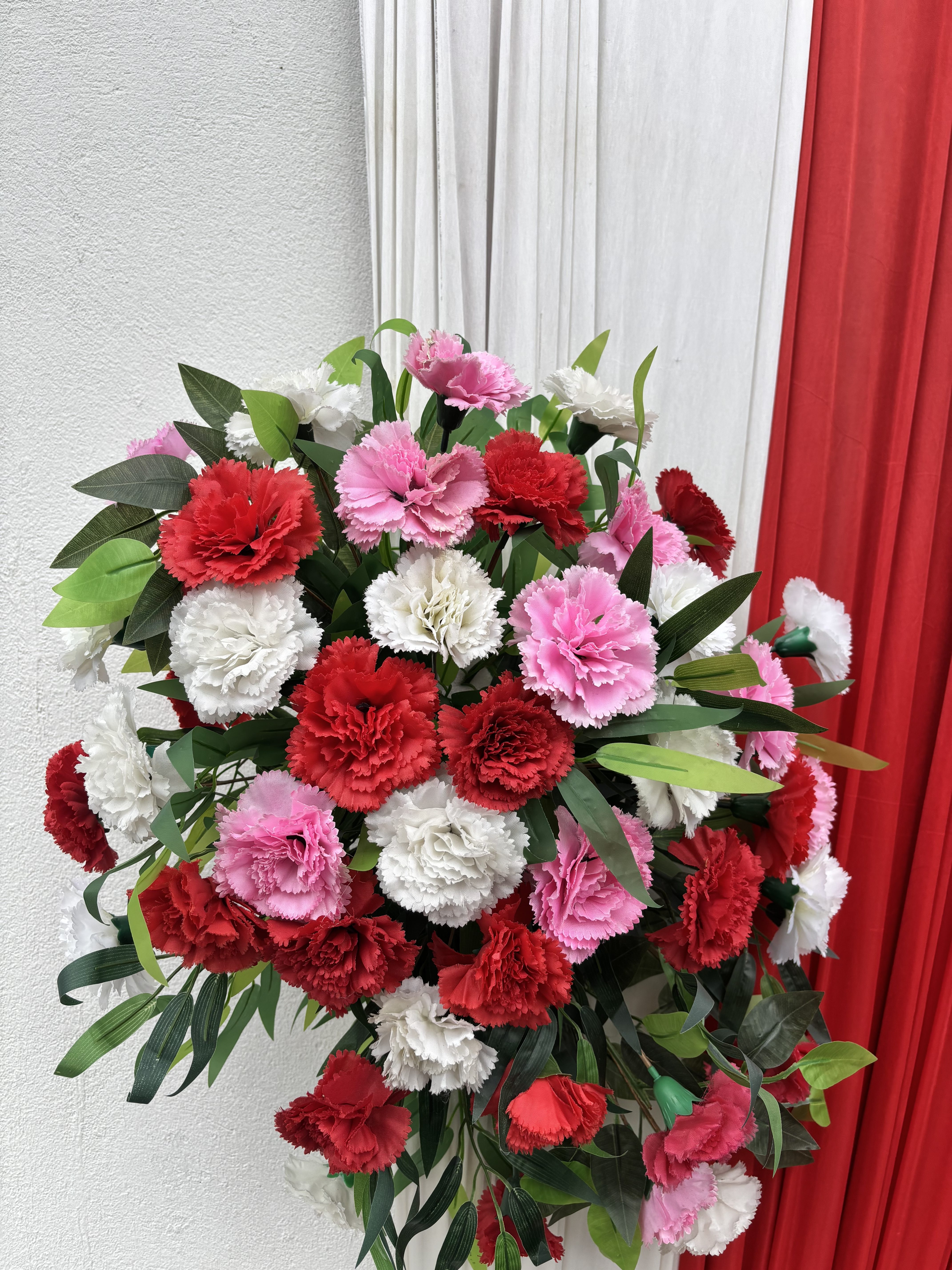 A vibrant bouquet of red, pink, and white carnations with green leaves against a backdrop of white and red fabric.