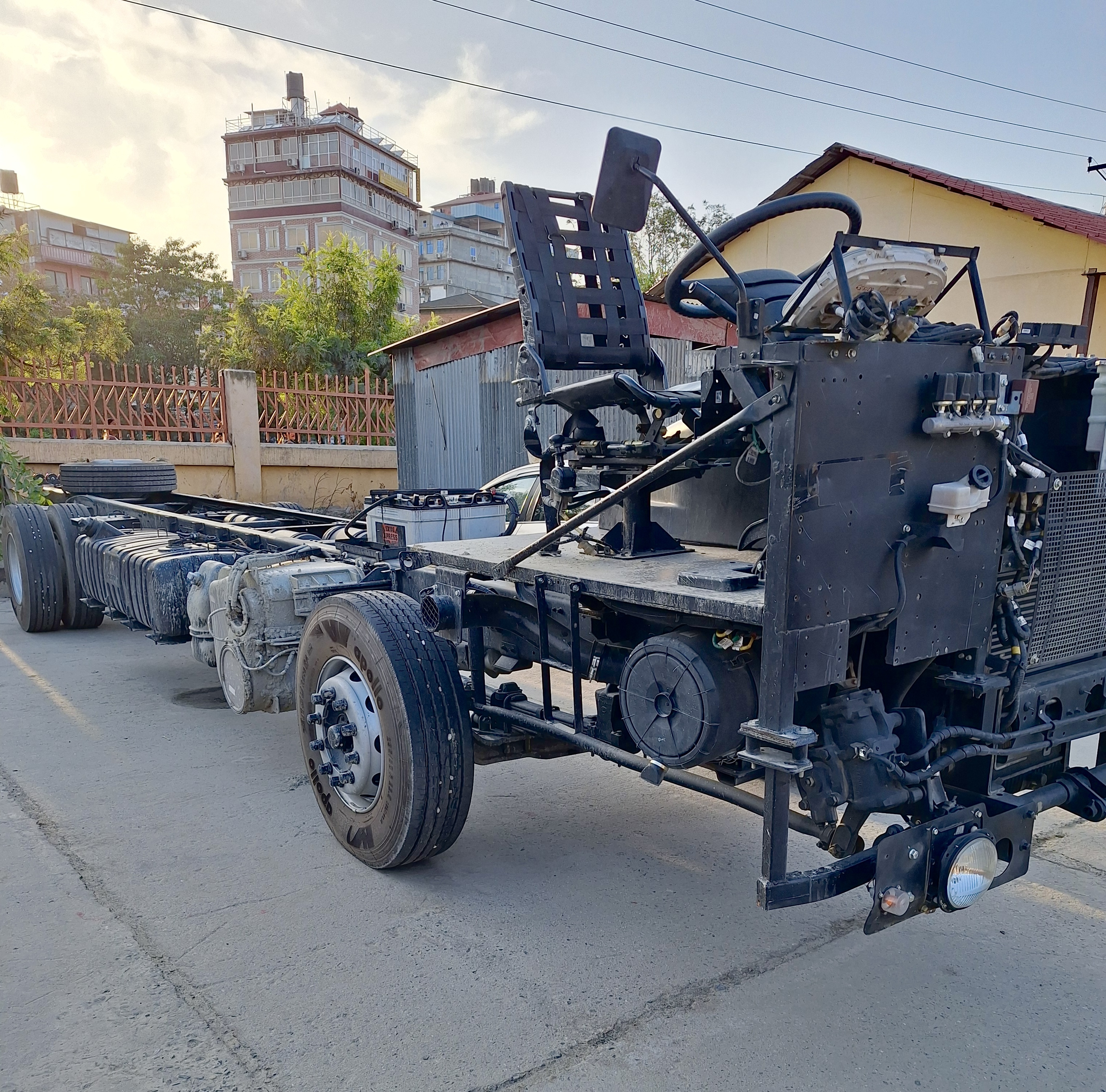 Raw unpainted heavy vehicle chassis parked outdoors on concrete, showing exposed mechanical components, open steering column, black driver seat, and heavy-duty axles.