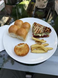 A white plate holds dinner rolls, a crispy hash brown, and a toasted sandwich filled with colorful vegetables and cheese.