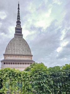 The famous Mole Antonelliana rises high into the cloudy sky, surrounded by green plants below. A striking landmark view captured in Turin (Torino), Italy.