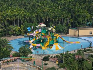Colorful water park playground with twisting slides and shallow pool, surrounded by tropical palm trees and lush greenery.
