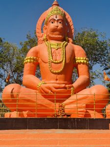 A serene, vibrant orange statue of Lord Hanuman sitting in a reflective pose, decorated with intricate golden accessories and framed against a bright, cloudless sky.