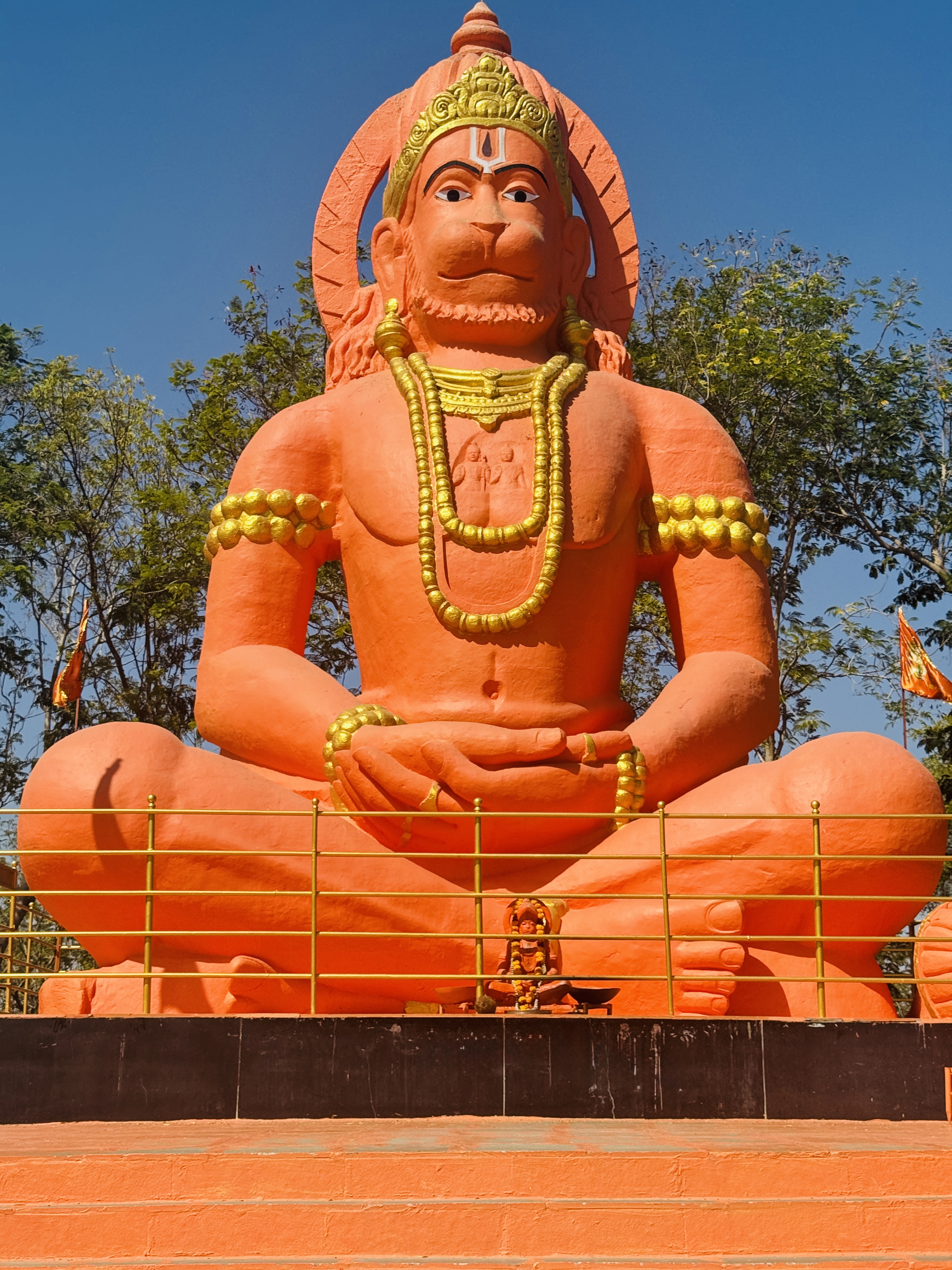 A serene, vibrant orange statue of Lord Hanuman sitting in a reflective pose, decorated with intricate golden accessories and framed against a bright, cloudless sky.