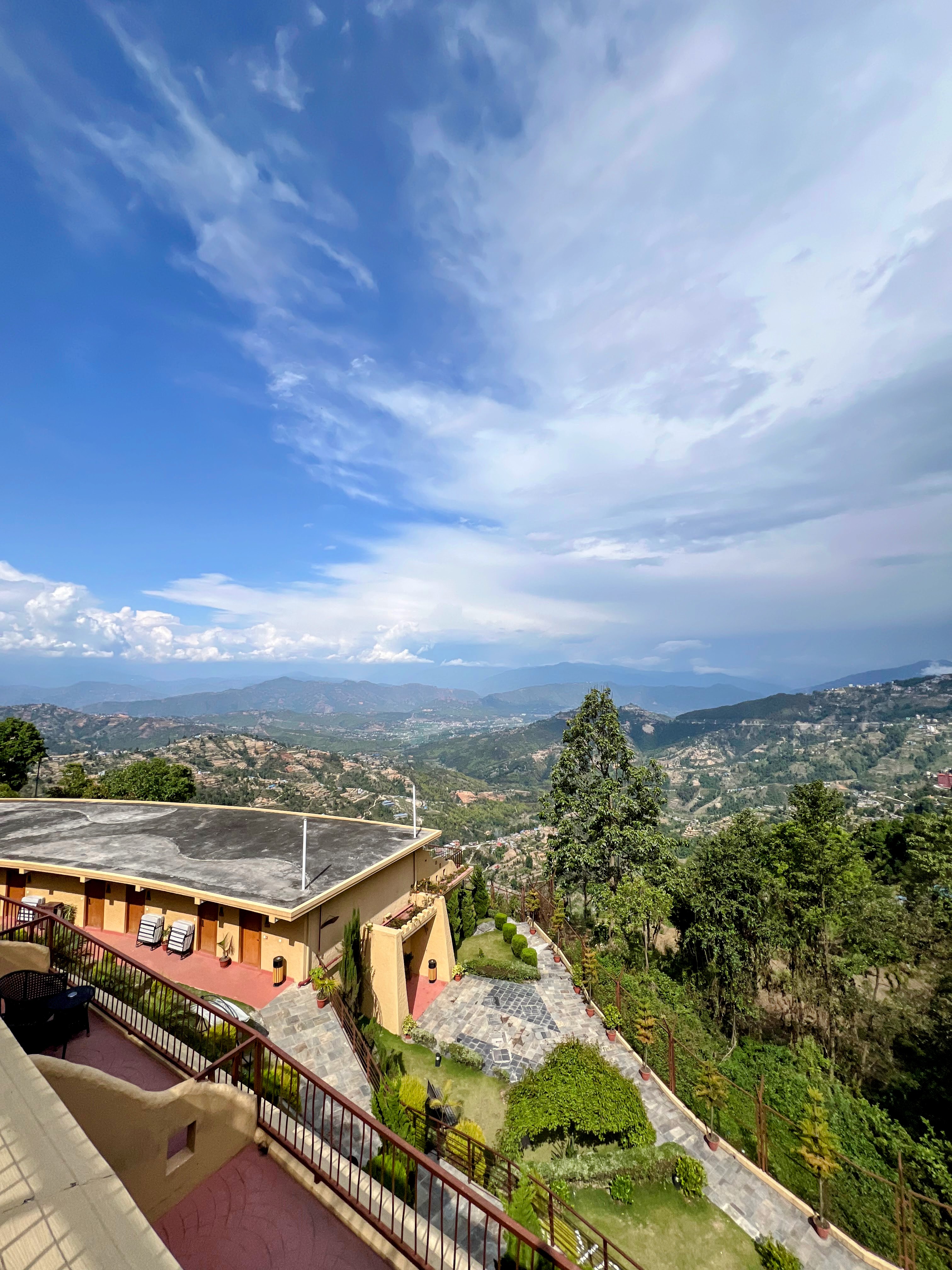 A high-angle, wide view of a building overlooking a vast, green hilly valley under a blue sky with scattered clouds.