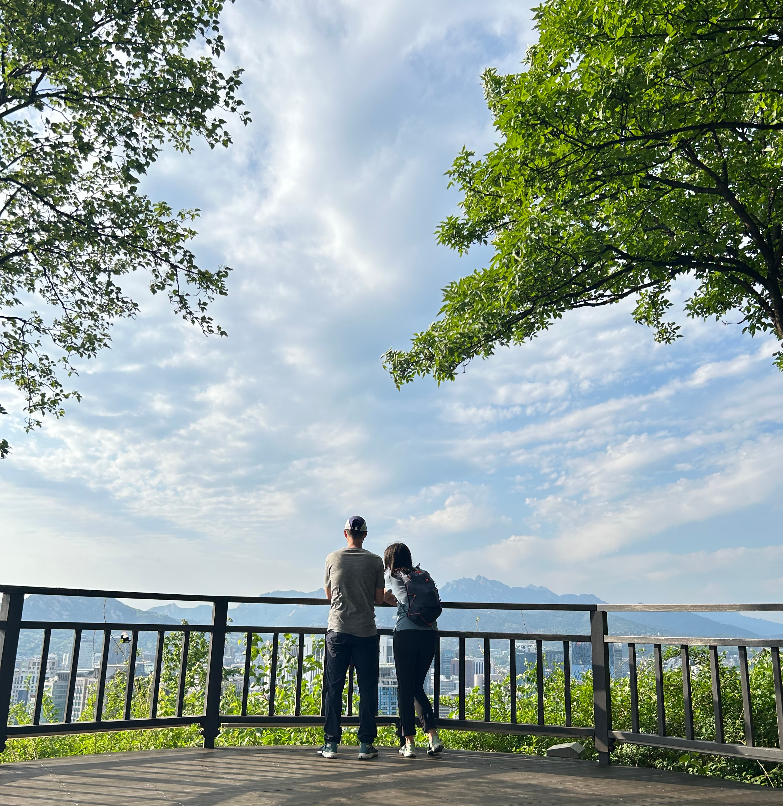 Two people stand at a wooden overlook, gazing at a cityscape under a bright blue sky. Trees frame the scene, creating a peaceful atmosphere.
