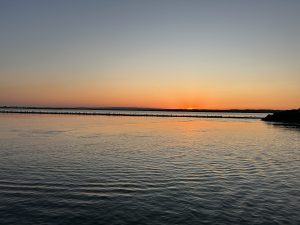 A serene scene captures a tranquil body of water at sunset. The sky transitions from a soft blue at the top to warm shades of orange and pink near the horizon, where the sun is dipping below the landscape. 