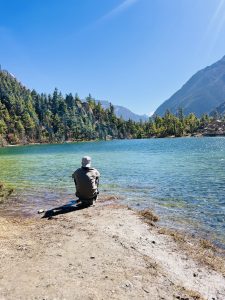 A person in outdoor clothing sits by a serene blue lake, surrounded by lush green trees and distant mountains under a clear, sunny sky.