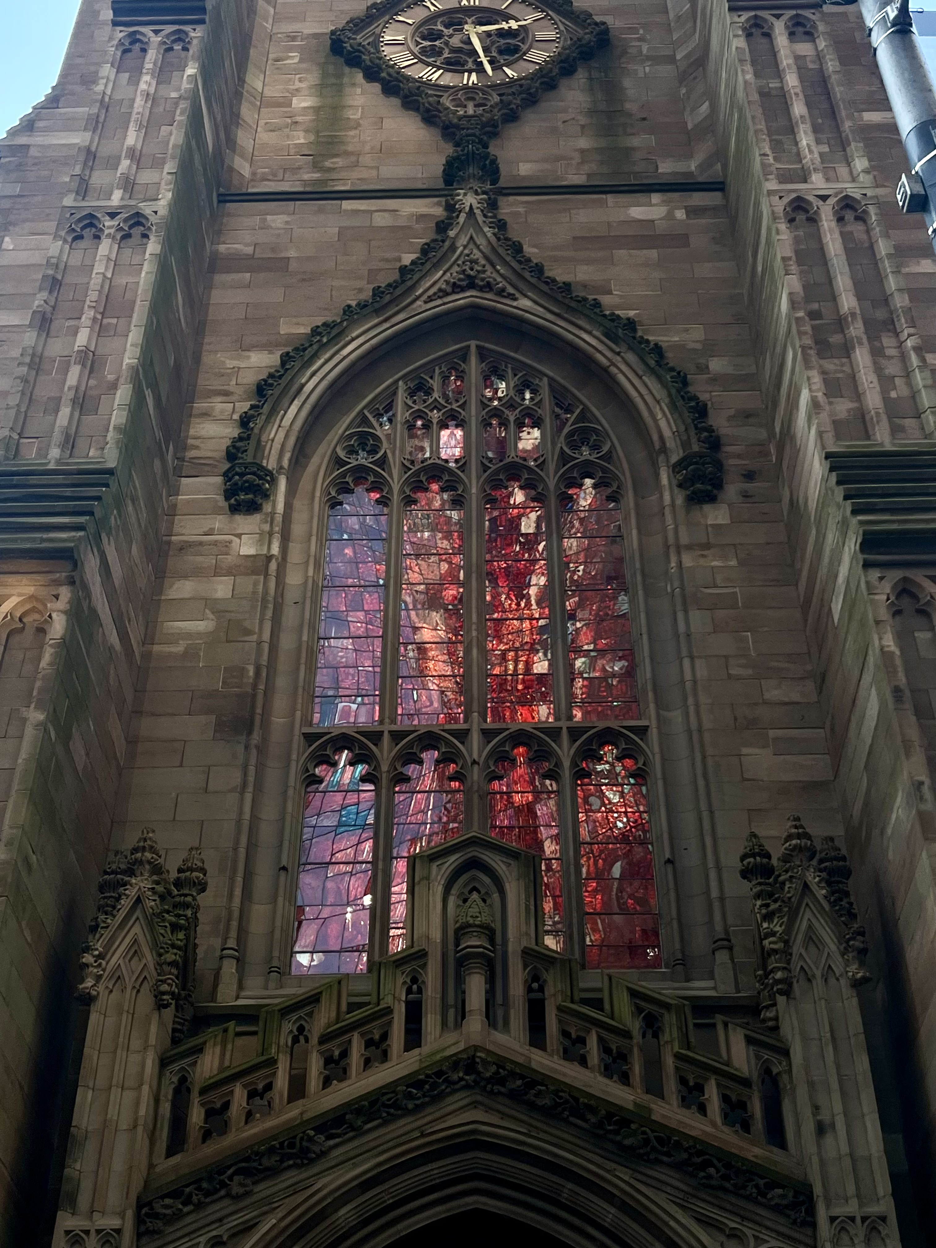 Upward view of a Gothic stone church facade featuring a large red stained glass window with ornate stone tracery and a diamond-shaped clock above it.