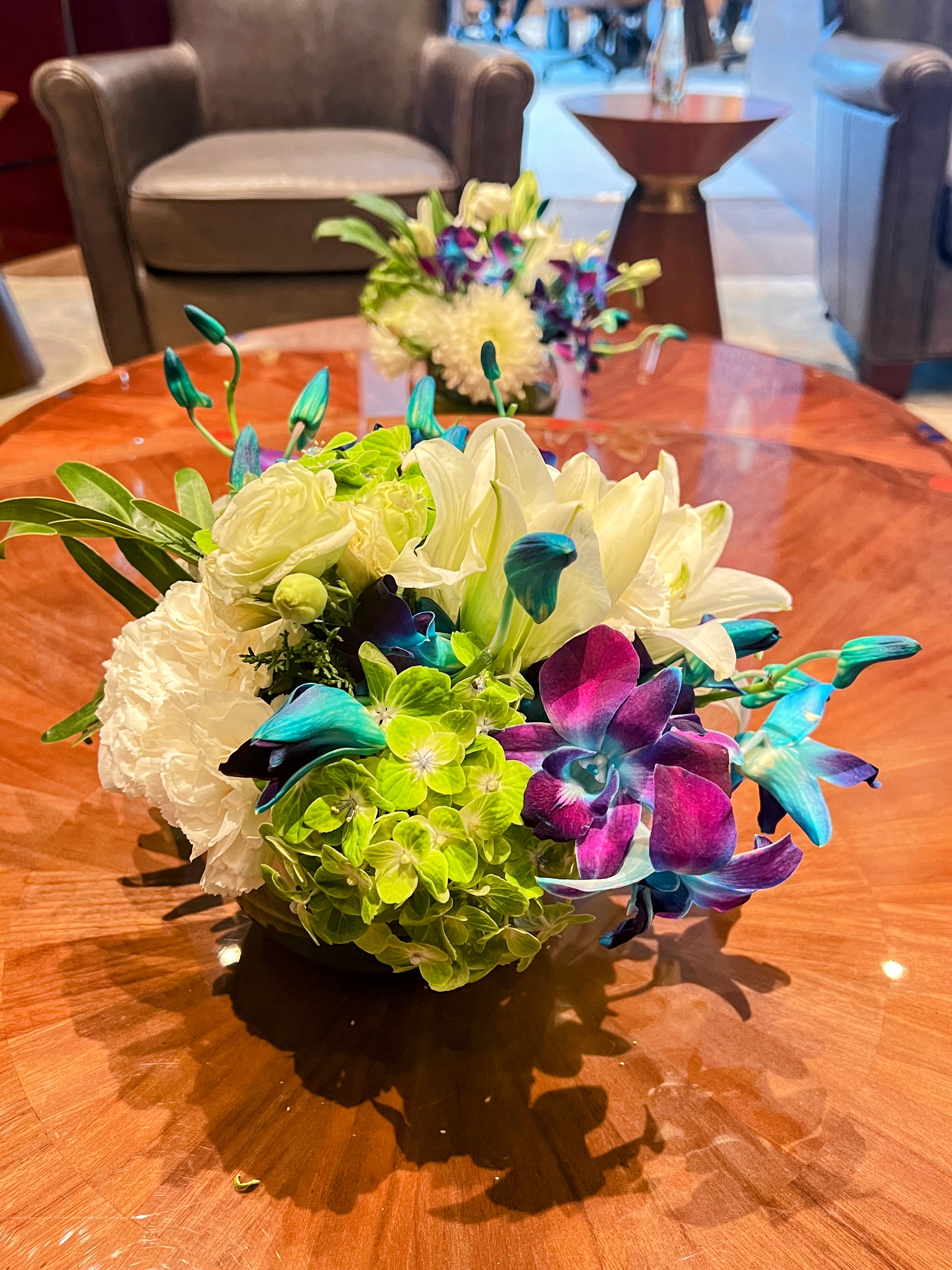 A vibrant floral arrangement featuring white chrysanthemums, green buds, and brilliant blue and purple orchids polished wooden table.