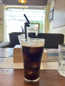 A glass of iced coffee with foam on top sits on a wooden table in a cozy cafe. The background shows soft lighting and a blurred view of furniture.