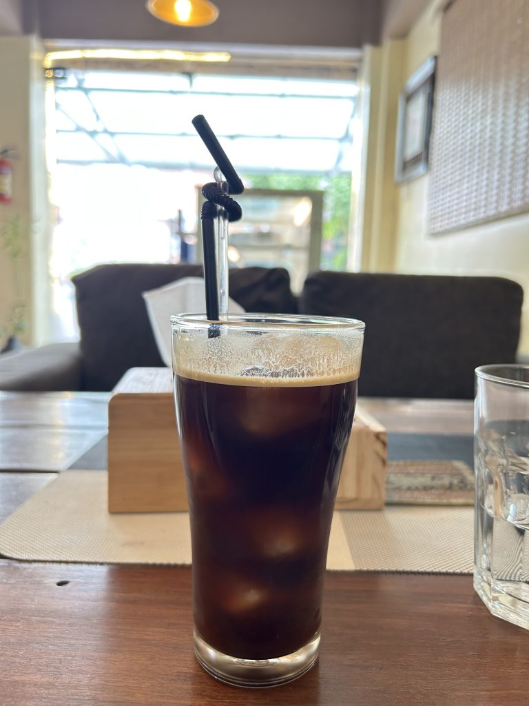 A glass of iced coffee with foam on top sits on a wooden table in a cozy cafe. The background shows soft lighting and a blurred view of furniture.