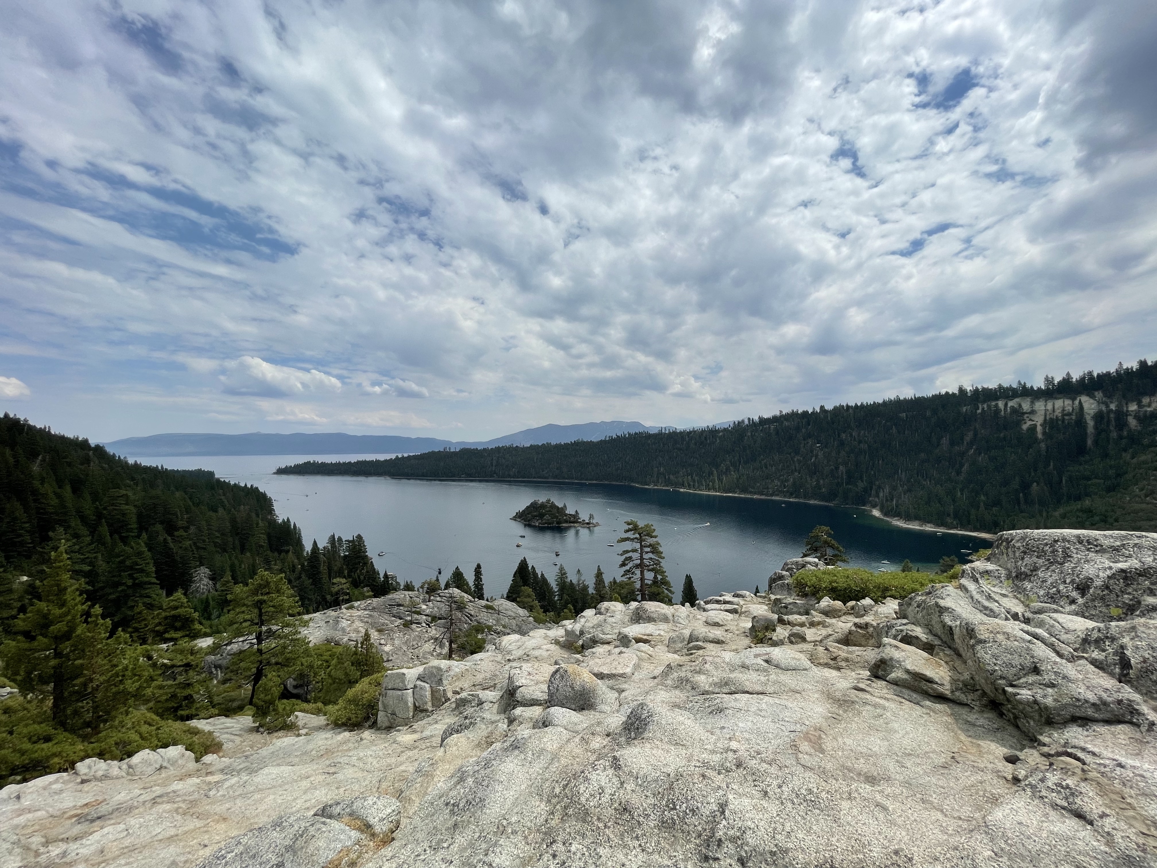 A high-angle panoramic vista of Emerald Bay at Lake Tahoe, taken from a rocky granite overlook. The small, pine-covered Fannette Island is visible in the center of the calm, dark blue bay, which is flanked by steep, heavily forested mountain ridges under an expansive sky filled with dramatic white and gray clouds.