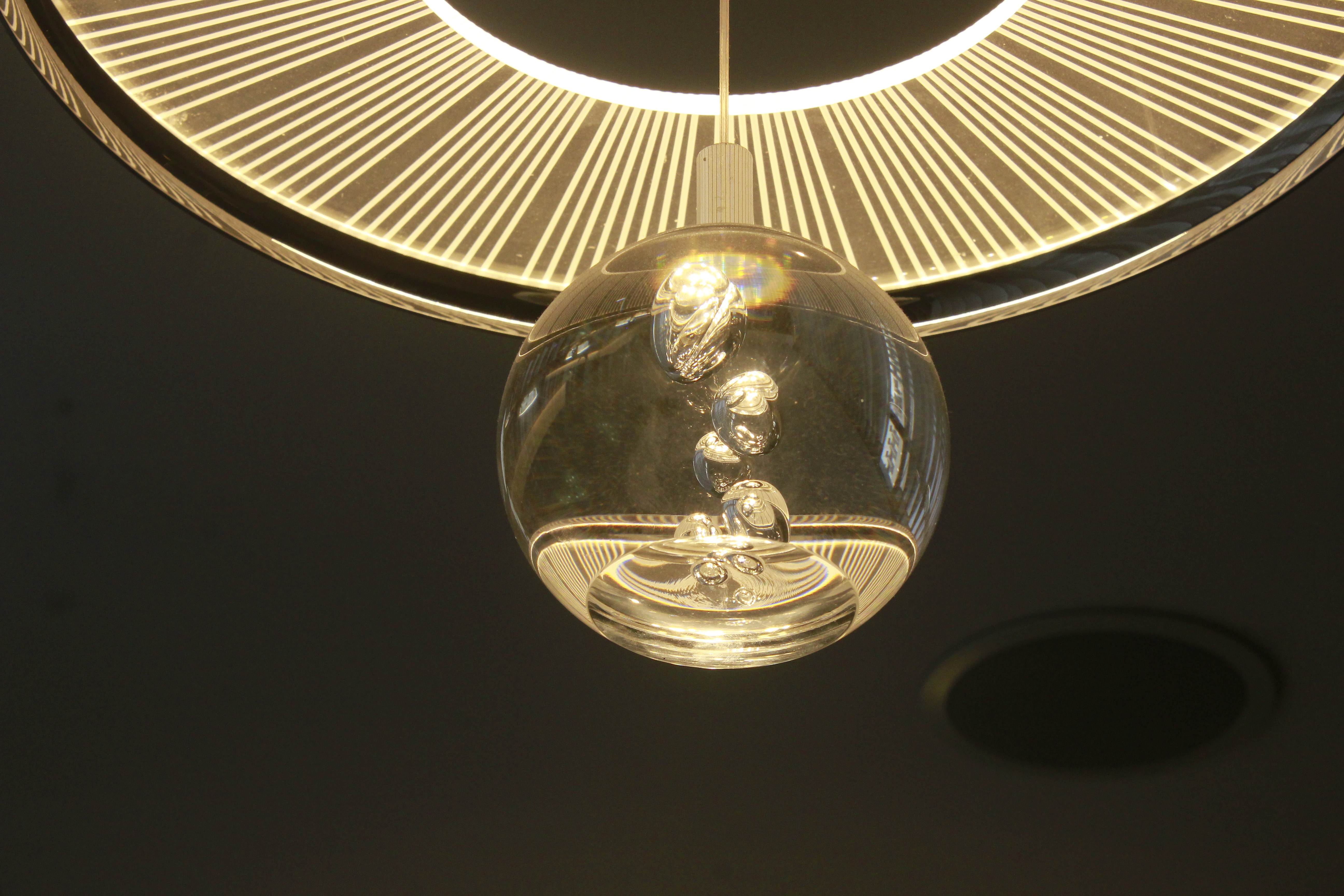 A clear glass globe pendant light hanging from a circular glowing ring light fixture against a dark ceiling, with reflective chrome spheres visible inside the glass ball.