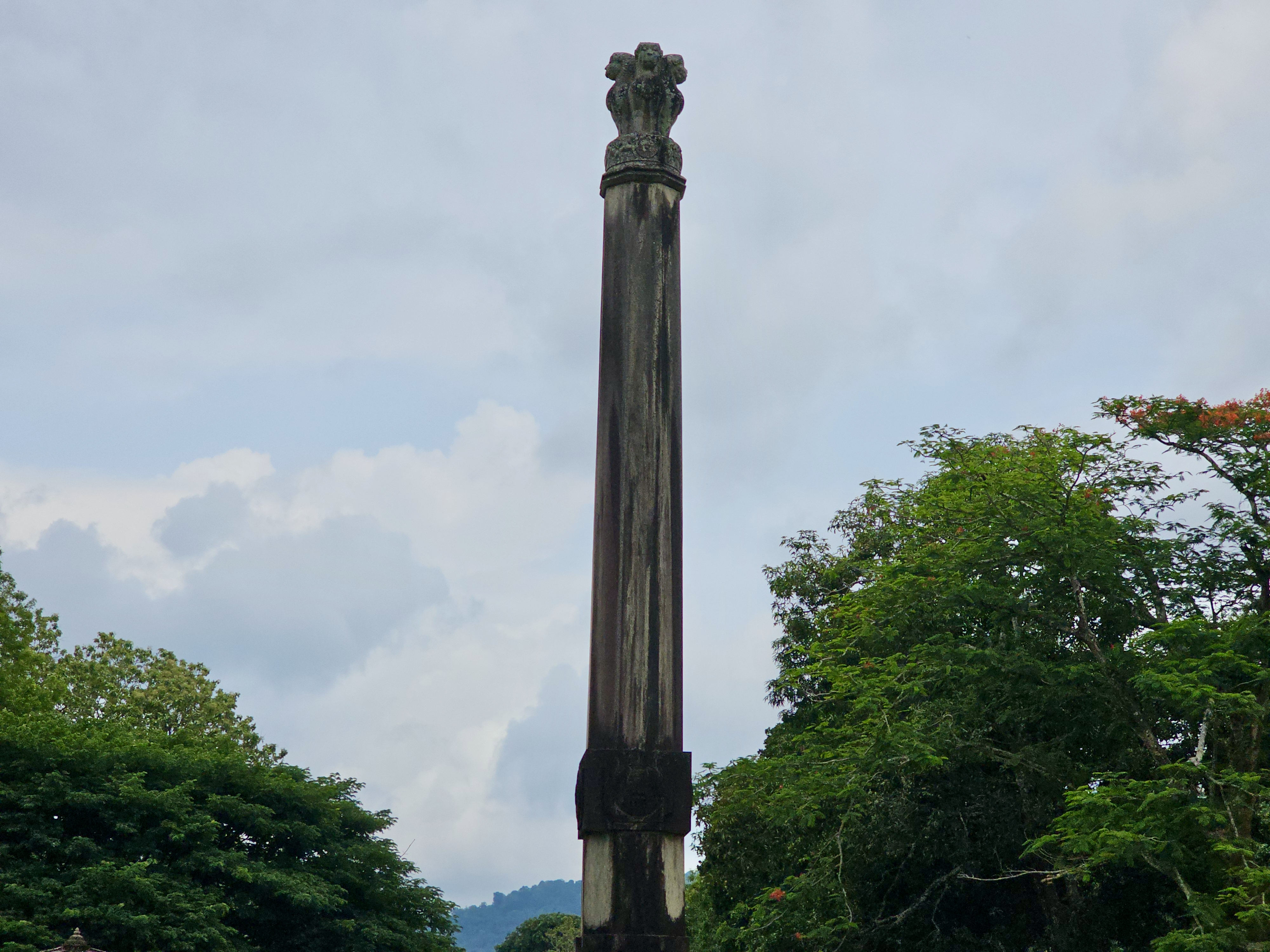 A tall, historic stone pillar standing proudly against a cloudy sky creates a calm, heritage-filled atmosphere. The greenery around the monument adds natural beauty to the scene. Captured from Parambikulam Tiger Reserve, Palakkad.
