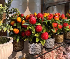 Pots of artificial plants with vivid red and yellow small fruit sit on a glass shelf. The pots have intricate black and gold patterns.