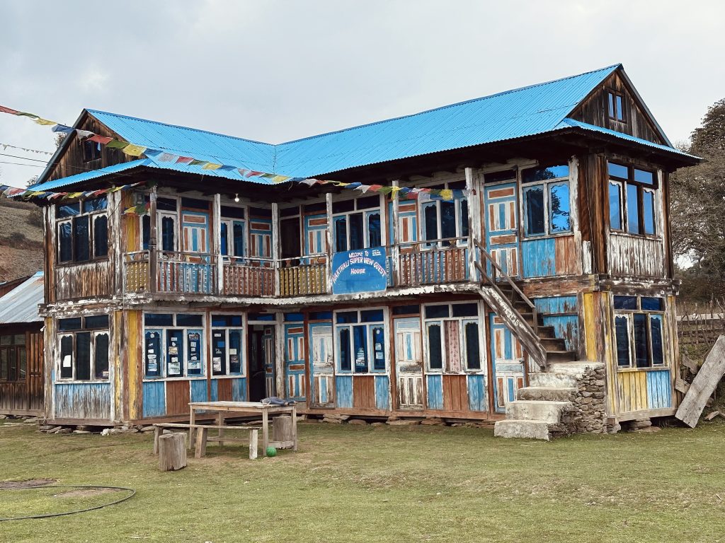 Rustic building with a blue roof, colorful window frames, a sign, and prayer flags adorning the structure.