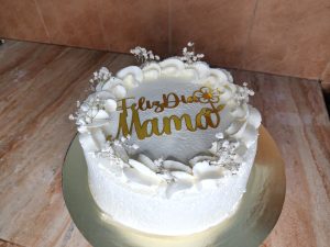 Cake decorated with white cream and delicate floral details, featuring an elegant golden topper that says “Feliz Día Mamá.” Its simple and sophisticated design conveys love, celebration, and a special atmosphere to honor mothers.
