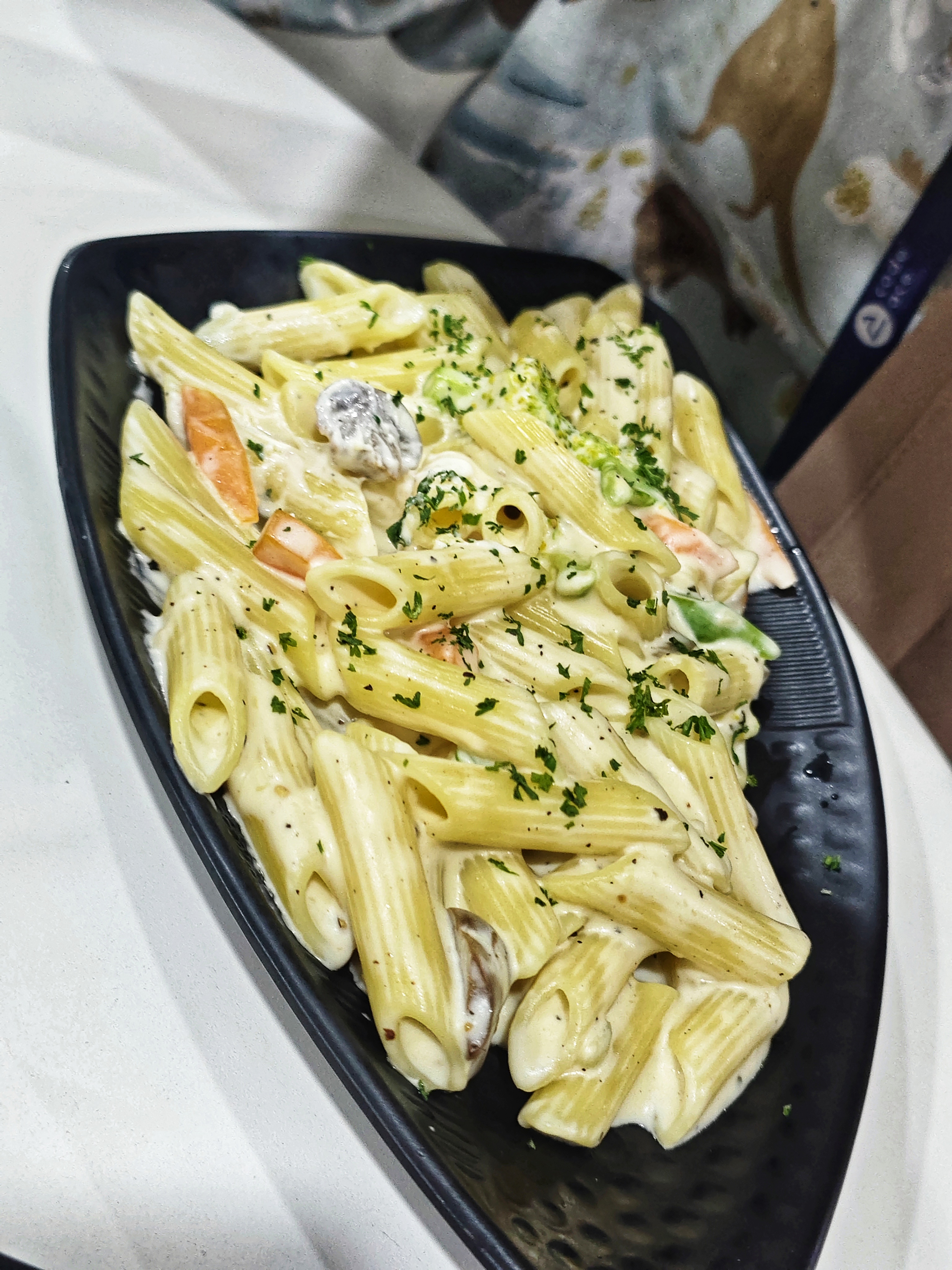A close-up view of a black oval plate containing creamy pasta, featuring penne shapes mixed with slices of vegetables, including pieces of orange carrots and green herbs sprinkled on top for garnish