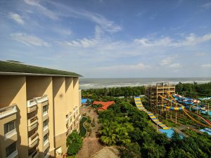 A seaside view featuring a multi-story beige building on the left, a green landscape with palm trees, and a colorful water park with slides on the right. The ocean and a partly cloudy sky create a serene background.