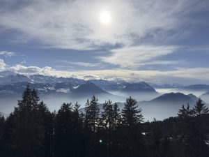 Snow-covered mountain range under a bright sun with dramatic swirling clouds, misty alpine valleys, and dark evergreen forest silhouettes in the foreground. Panoramic winter landscape featuring rugged peaks, blue sky, atmospheric haze, and serene natural scenery. High-altitude mountain vista in the Swiss Alps with sunlight breaking through.
