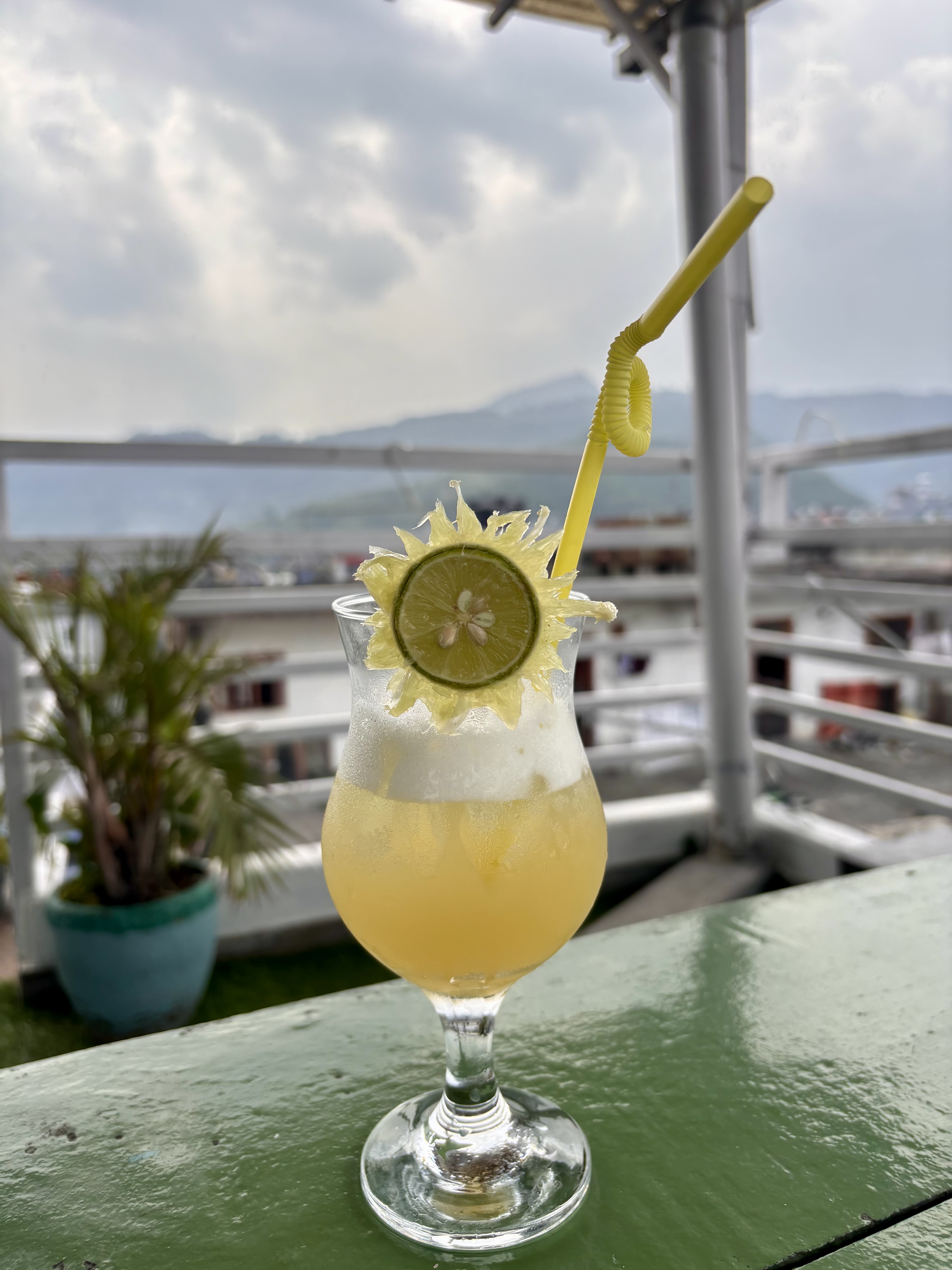 A tropical drink garnished with a lime slice and yellow straw sits on a green table, with a hazy mountain view in the background.