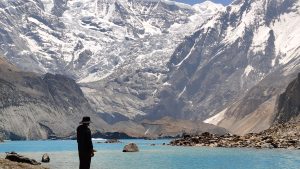 A person in dark clothing and a hat stands by a turquoise lake, surrounded by snow-capped mountains under a clear blue sky.