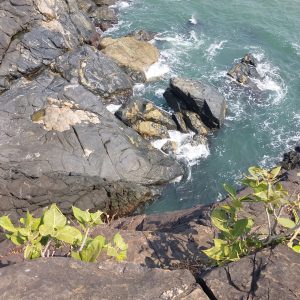 The image shows a rocky coastline where dark, jagged rocks meet the turquoise water of the ocean. There are patches of light-colored stones scattered among the darker ones.