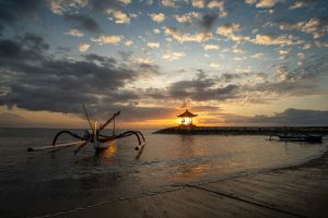 Sunrise at Sanur beach, Bali, Indonesia. The sun is behind a gazebo, which makes it look somewhat like a lantern. A traditional wooden boat lies on the beach. The sky is partially cloudy.
